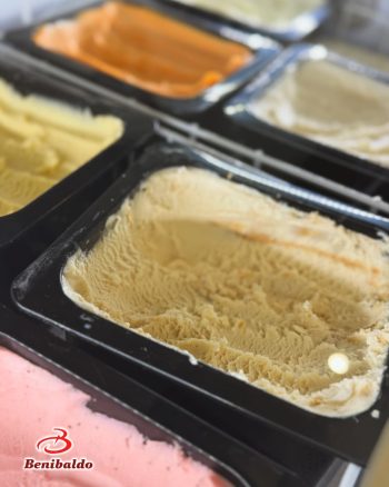 Pale beige gelato in a black display tub at a gelato shop, with other flavors (orange, yellow, pink) visible in the background; Benibaldo logo bottom left.