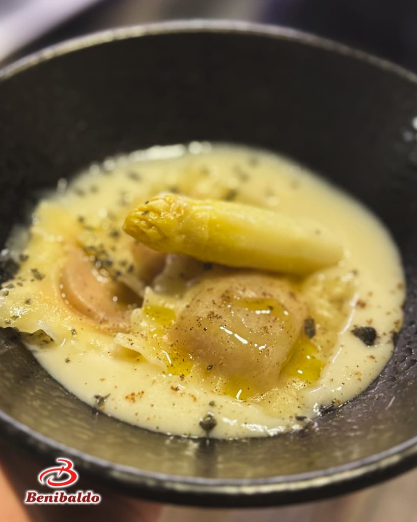 Creamy white sauce with pasta pieces simmering in a black skillet, lightly garnished with herbs. A yellow vegetable piece rests on top. Brand watermark at corner.