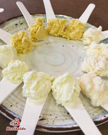Assorted cheese samples on white tasting spoons arranged around a plate, with the Benibaldo logo visible in the corner.