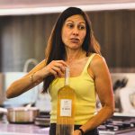 Woman in a yellow tank top in a kitchen holding a tall bottle of white wine labeled Tres Maris.