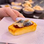 Canapé: small toasted bread topped with anchovy on jam, placed on a white plate as a hand nears it; catering bowls in soft focus in the background.