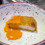 Slice of cheesecake with a crumb crust on a speckled ceramic bowl, topped with orange sauce on the side.