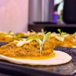 Tortilla topped with shredded meat in sauce and a microgreen garnish on a dark plate, close-up.