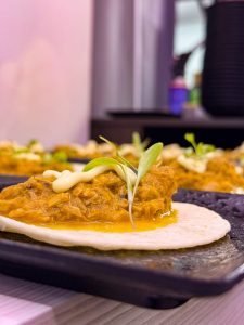 Tortilla topped with shredded meat in sauce and a microgreen garnish on a dark plate, close-up.
