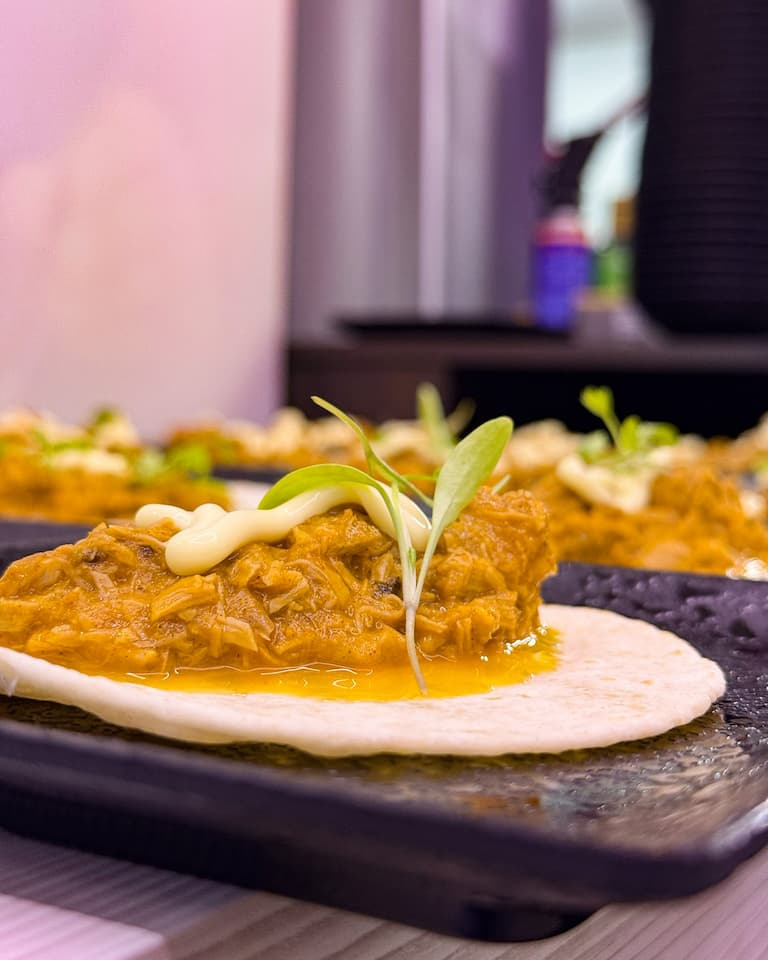 Tortilla topped with shredded meat in sauce and a microgreen garnish on a dark plate, close-up.