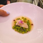Fine-dining plate: white fish on a bed of bright green peas in a light broth, garnished with a purple flower.