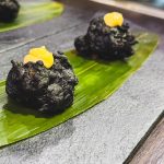 Two black fried morsels topped with a dollop of orange sauce on a green leaf, presented on a slate surface