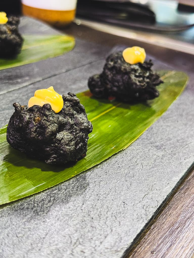 Two black fried morsels topped with a dollop of orange sauce on a green leaf, presented on a slate surface