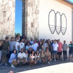 Large group photo outside a stone building with abstract metal wall art; Benibaldo logo bottom left.