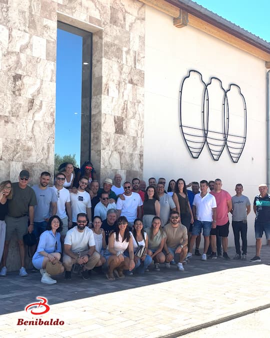 Large group photo outside a stone building with abstract metal wall art; Benibaldo logo bottom left.