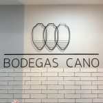 Three overlapping wireframe wine glasses logo above the text 'BODEGAS CANO' on a white wall with brick subway tiles; Benibaldo logo bottom-left.