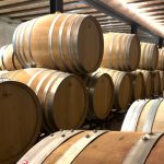 Rows of wooden wine barrels stacked on racks in a dimly lit aging cellar, Benibaldo logo visible in the corner.