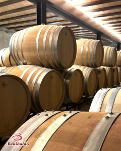 Rows of wooden wine barrels stacked on racks in a dimly lit aging cellar, Benibaldo logo visible in the corner.