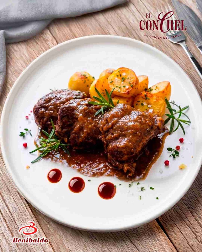 Plate with braised beef in brown sauce, roasted potatoes, and rosemary on a white dish; garnished with herbs and red peppercorns, on a wooden table (logos in corners).