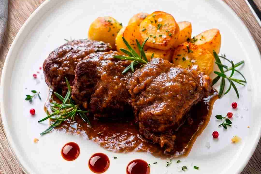 Plate with braised beef in brown sauce, roasted potatoes, and rosemary on a white dish; garnished with herbs and red peppercorns, on a wooden table (logos in corners).