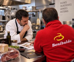 Two chefs study a cookbook at a stainless steel kitchen counter, discussing a recipe with ingredients laid out nearby